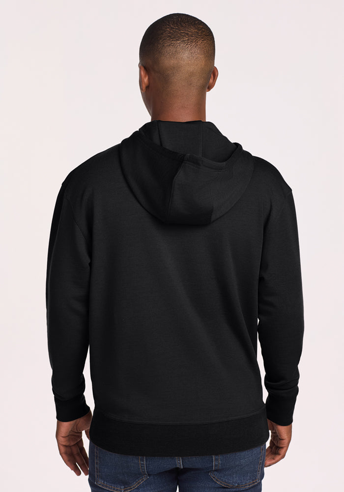 A man wears a Woolx Chase Hoodie in black, paired with blue jeans, standing with his back to the camera against a plain light background.