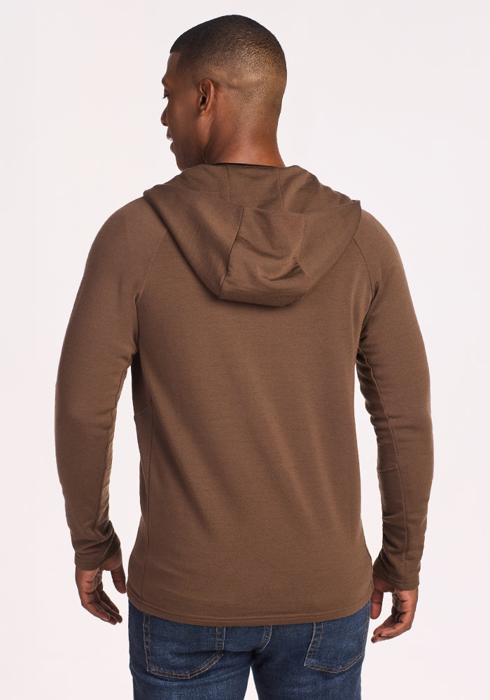 A man stands with his back to the camera, wearing the Woolx Boulder Hoodie in Bourbon Oak and blue jeans against a plain light background.