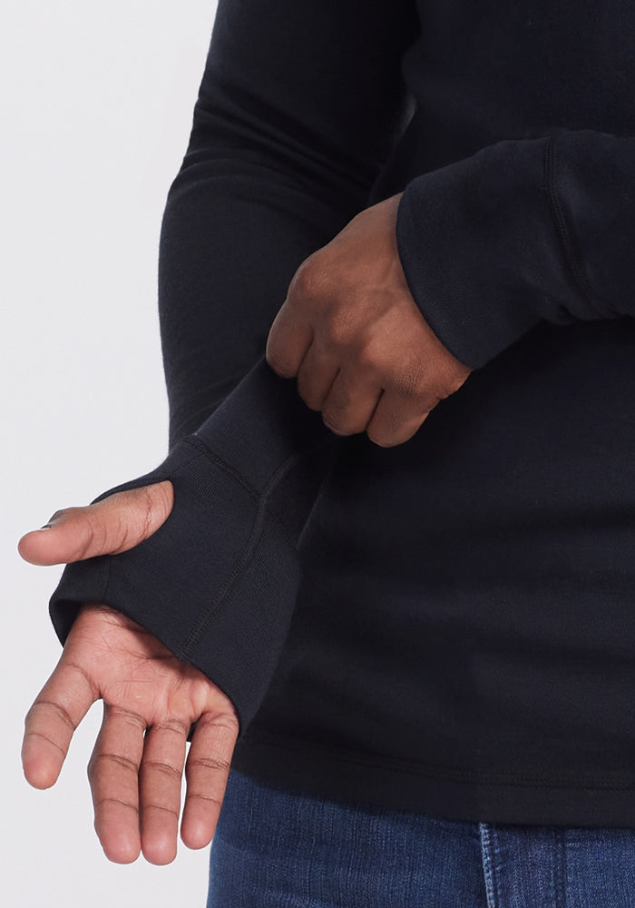 A person wears the Woolx Glacier Long Sleeve Crew in black, featuring thumb holes and pulled sleeves over their hand. Paired with blue jeans, it's perfect for chilly days. The background is plain and light-colored.