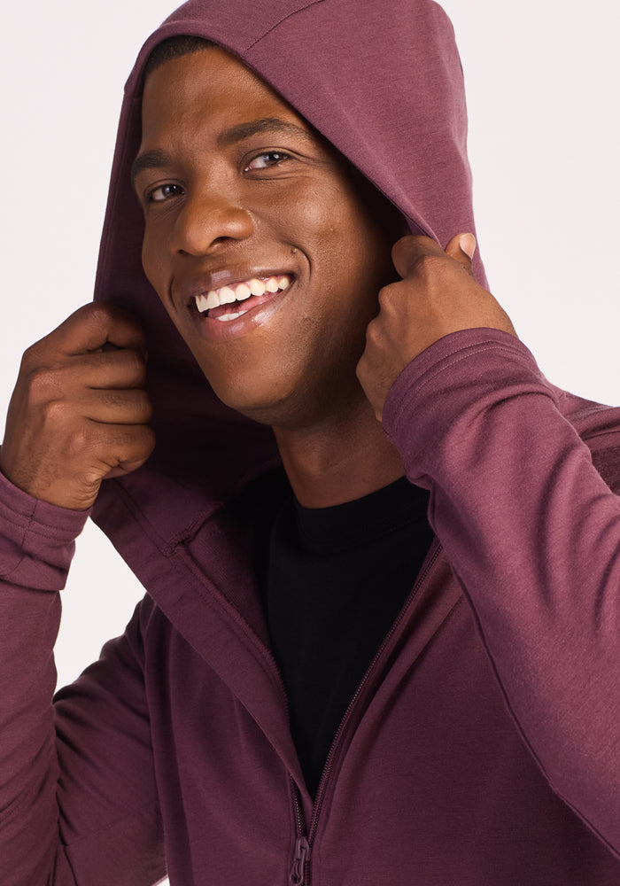 A smiling man wears the Woolx Boulder Hoodie in Grape Harvest, pulling the hood over his head with both hands. He has a black shirt underneath and stands against a light plain background.