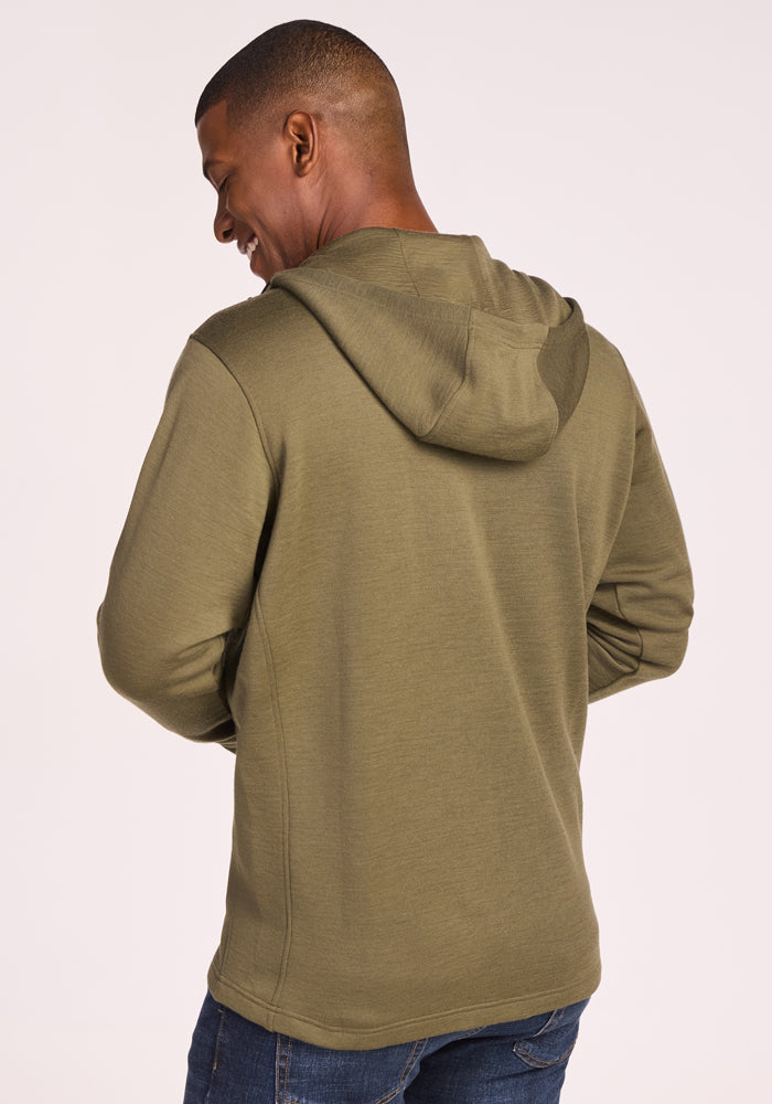 A man is smiling and facing away from the camera, wearing a Woolx Grizzly Sweatshirt in Winter Moss and blue jeans against a plain light background.