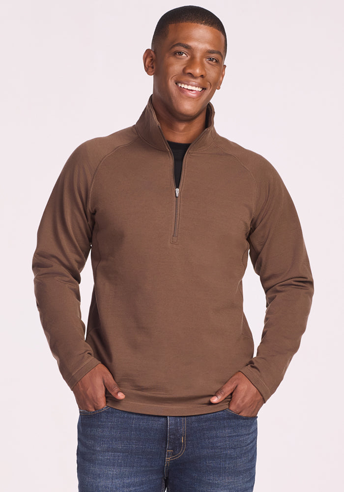 A smiling man wears the Woolx Blizzard 1/4 Zip in Bourbon Oak with blue jeans, standing with hands in his pockets against a plain light background. 