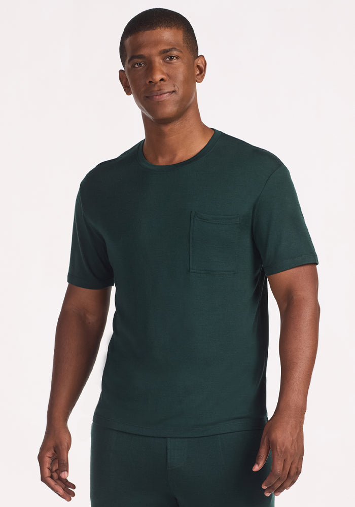 A man stands against a plain light background, wearing the Woolx Finn Top in Deep Timber—a dark green, temperature-regulating T-shirt with a chest pocket, ideal for men's pajamas. He looks forward neutrally with his arms relaxed at his sides. 
