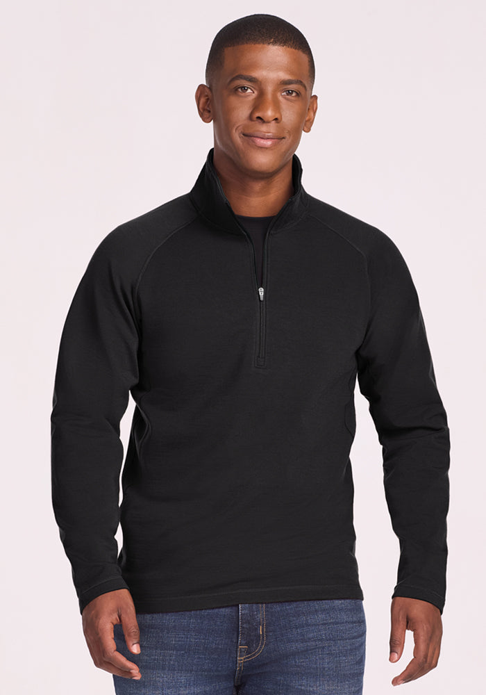 A man wears the Woolx Blizzard 1/4 Zip in black with blue jeans, standing against a light background and smiling gently with his hands relaxed at his sides. 