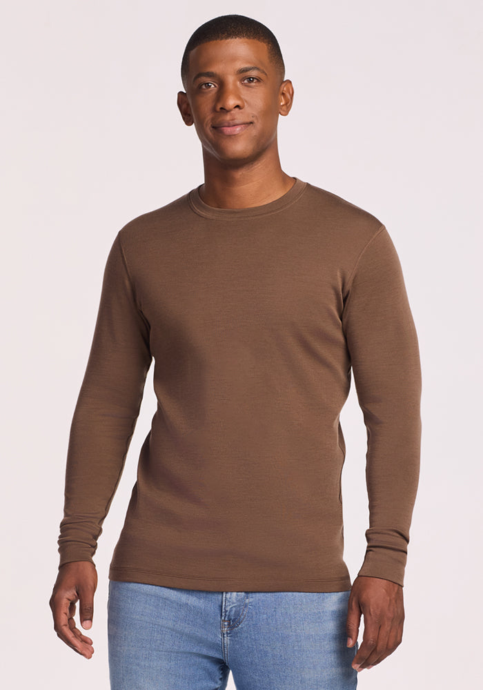A man stands facing forward, smiling slightly, against a plain light background, wearing the Woolx Explorer Base Layer in Bourbon Oak and blue jeans. 
