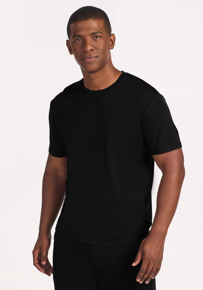 A man with short hair stands against a white background, smiling slightly while wearing the Woolx Finn Top in black—a plain, moisture-managing short-sleeve t-shirt featuring a chest pocket. 