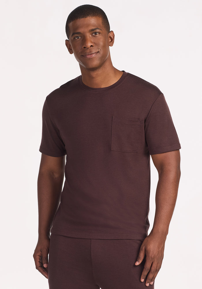 A man wears the Woolx Finn Top in Chocolate Plum—a short-sleeve, temperature-regulating t-shirt with a chest pocket—standing against a white background, smiling softly with one hand by his side. 