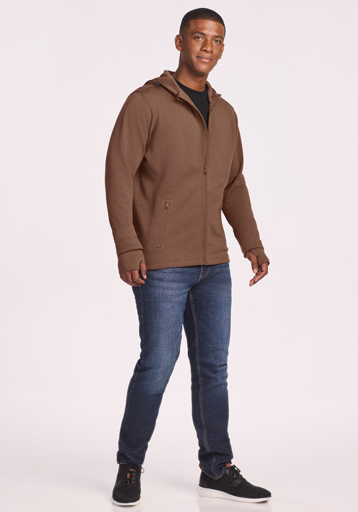 A man smiles, wearing the Woolx Grizzly Sweatshirt in Bourbon Oak with blue jeans and black sneakers, standing against a plain white background.