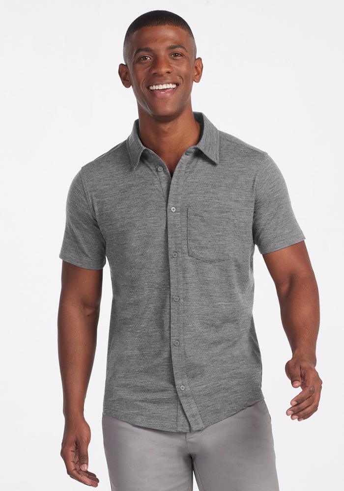 A smiling man wears the Woolx Endi Full Button Down Shirt in Graphite Heather, paired with light gray pants, standing against a plain white background.