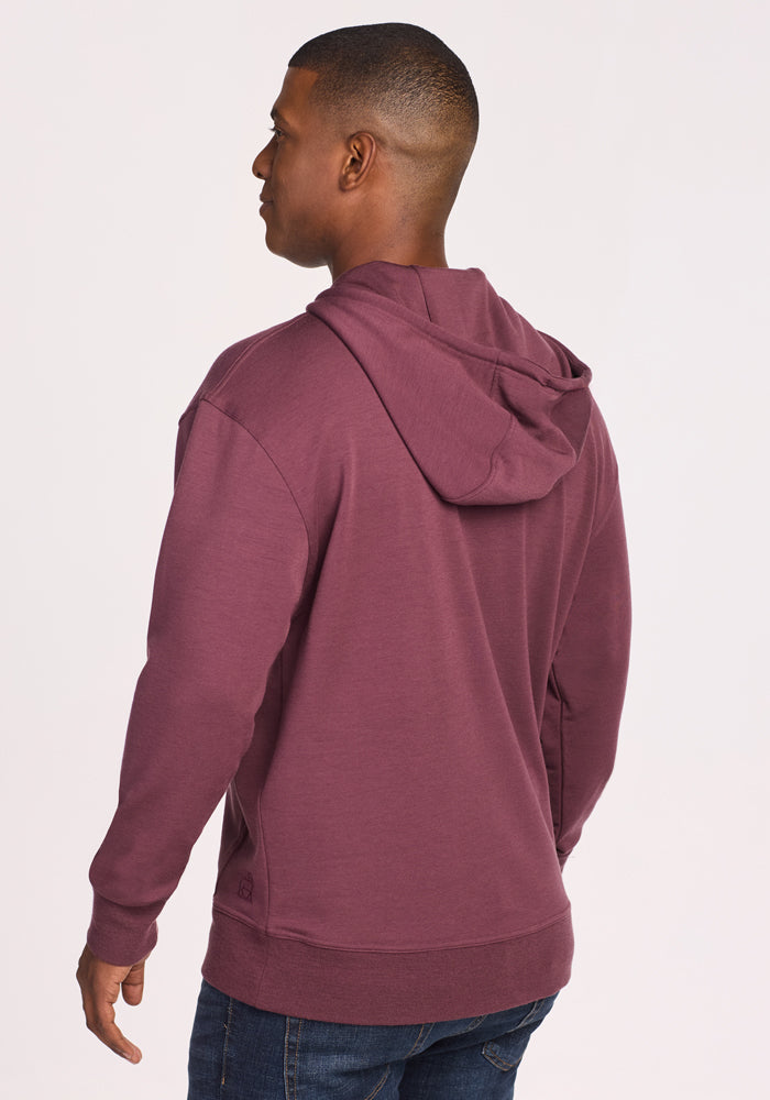 A man wearing a Woolx Chase Hoodie in Grape Harvest and blue jeans stands facing away from the camera, displaying the back and side of the hoodie against a plain light background.