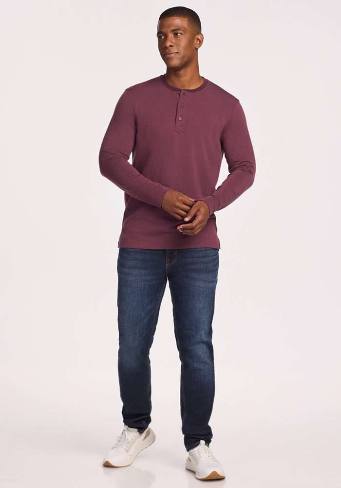 A man stands against a plain light background wearing the Woolx Jackson Henley in Grape Harvest, dark blue jeans, and white sneakers. He looks slightly to the side with his hands loosely clasped.