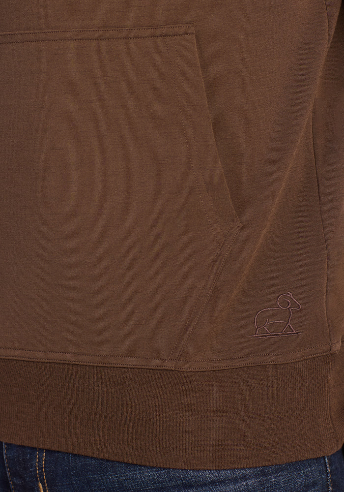 Close-up of the Woolx Chase Hoodie - Bourbon Oak, showing the front pocket and a small outlined elephant logo stitched near the ribbed hem. Blue denim jeans are partially visible below.