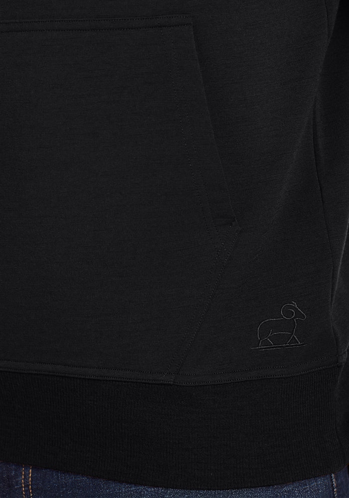 Close-up of the Woolx Chase Hoodie in black, showing the men’s wool hooded sweatshirt pocket with subtle stitching and a small minimalist elephant outline embroidered near it. Blue jeans are partially visible at the bottom.