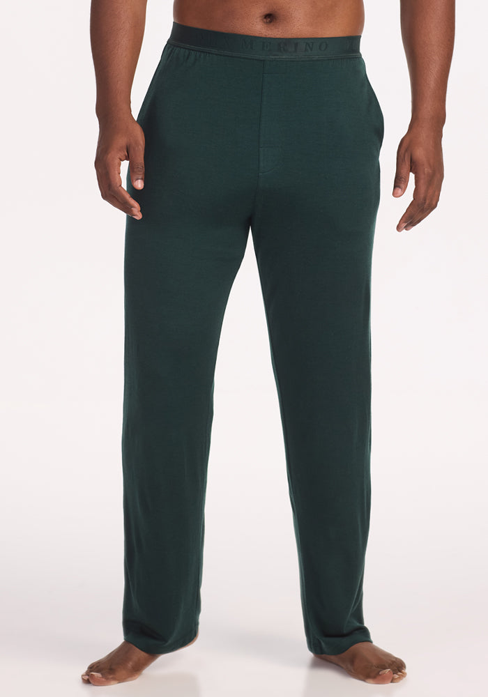 A person is barefoot, wearing Woolx Arlo Lounge Pants in Deep Timber, featuring Feathertouch fabric, an elastic waistband, and side pockets, standing against a plain white background. 