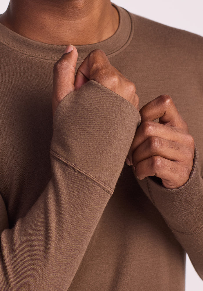 Wearing the Woolx Glacier Long Sleeve Crew in Bourbon Oak, a person pulls the sleeve over one hand, showcasing the rich fabric texture up close. Only their upper torso and hands are visible.
