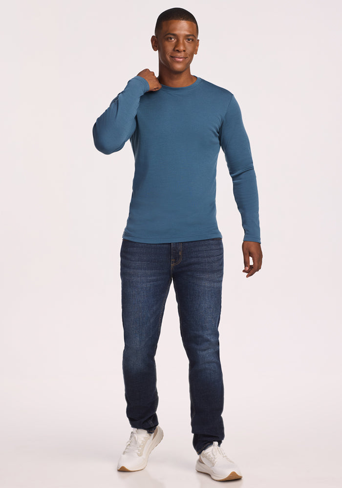 A man smiles against a plain background, wearing the Woolx Easton Crew Neck Shirt in Steel Blue, paired with dark blue jeans and white sneakers. One hand is raised to his neck, the other relaxed by his side.