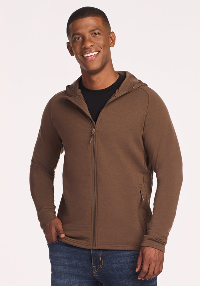 A smiling man with short hair wears the Woolx Boulder Hoodie in Bourbon Oak over a black shirt and blue jeans, standing with one hand in his pocket against a plain light background. 