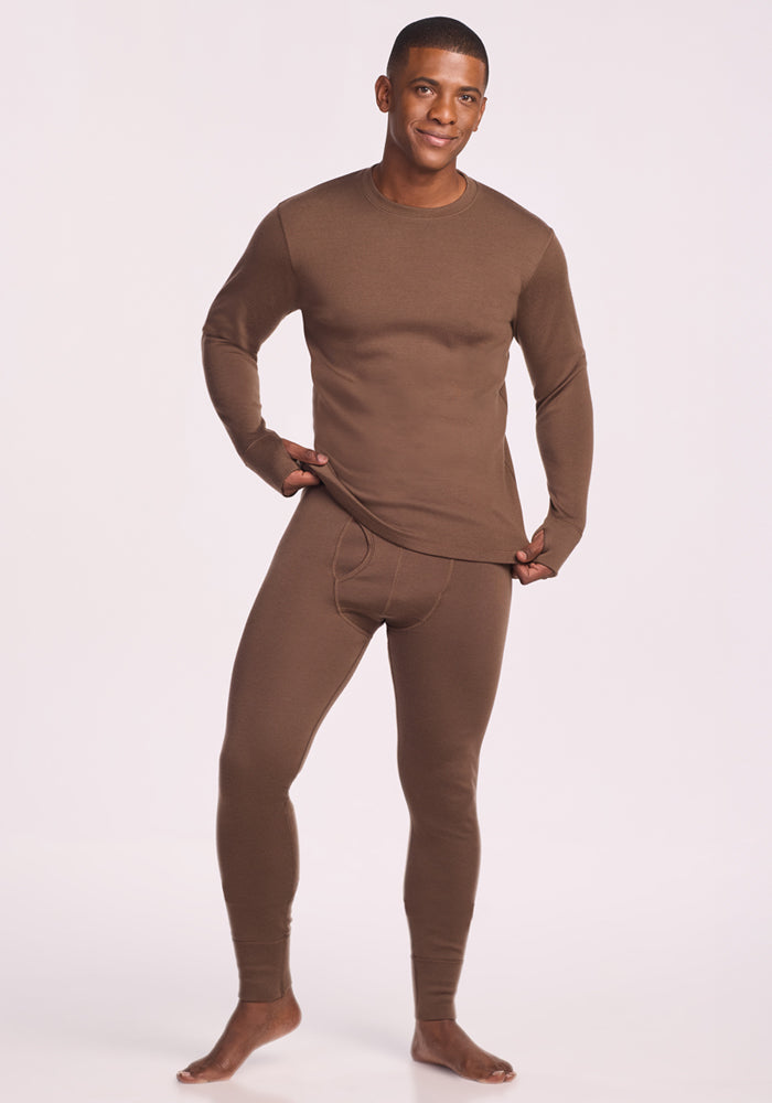 A man stands barefoot against a plain backdrop, smiling as he wears the Woolx Glacier Long Sleeve Crew in Bourbon Oak. He lifts the hem to show off its merino wool softness and odor-resistant comfort.