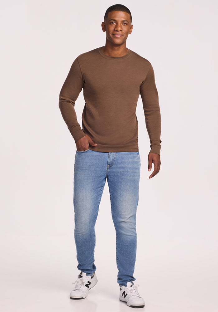 A man smiles against a plain white background, wearing the Woolx Explorer Base Layer - Bourbon Oak, light blue jeans, and white sneakers with "N" logos.