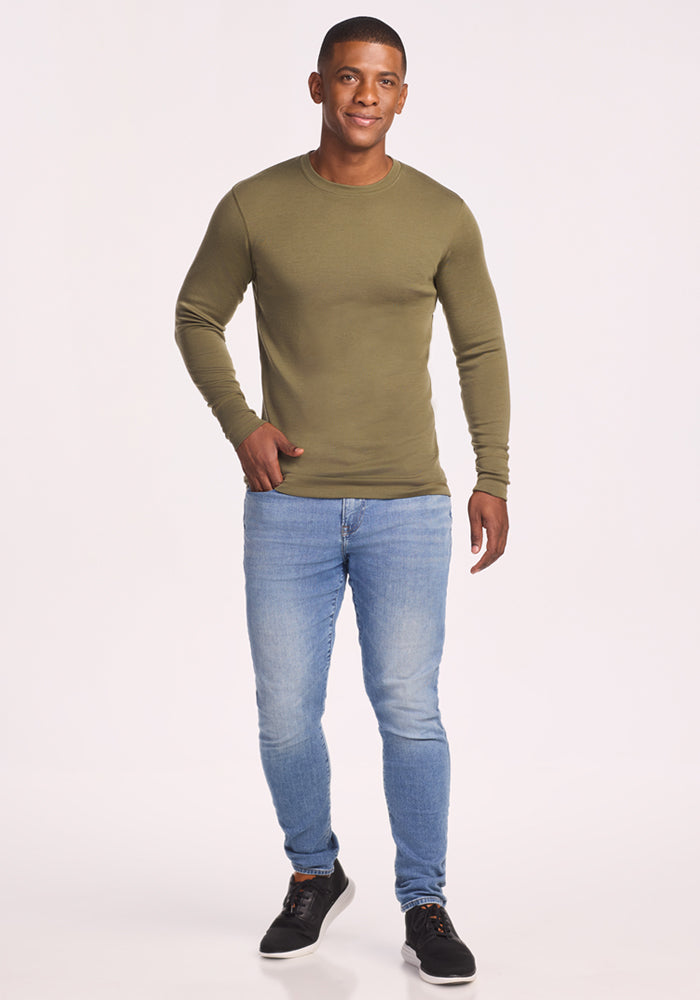 A man smiles, wearing the Woolx Explorer Base Layer in Winter Moss, paired with light blue jeans and black sneakers, standing against a plain white background.