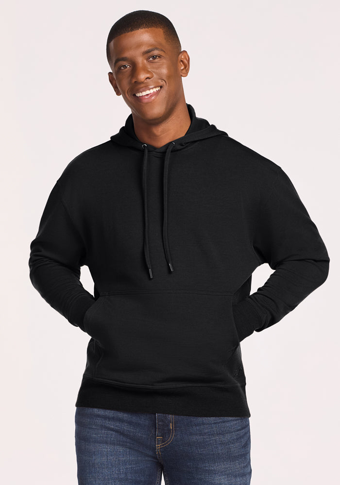 A young man smiles wearing the Woolx Chase Hoodie - Black, featuring moisture-wicking fabric and a front pocket, paired with blue jeans against a simple backdrop. 