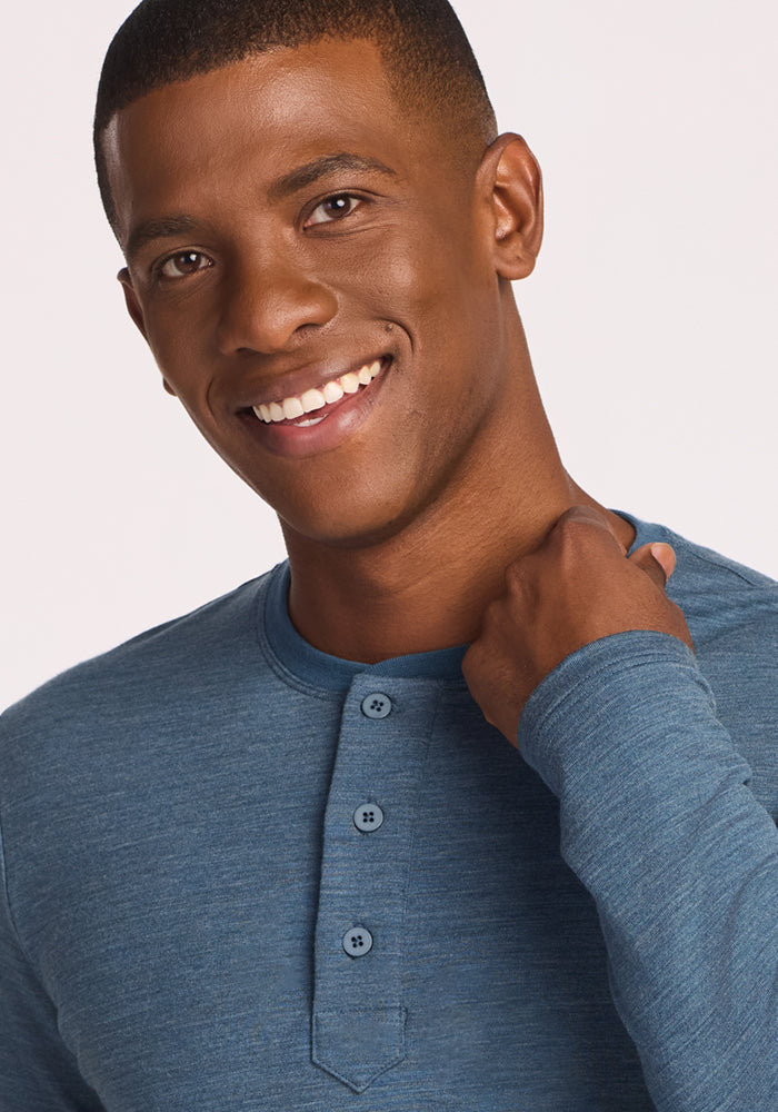 A man wearing the Woolx Jackson Henley in Steel Blue smiles warmly at the camera, holding his shirt collar with one hand against a plain, light-colored background.