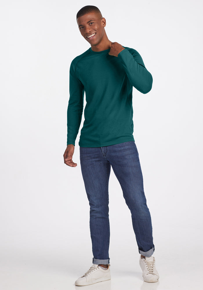 A smiling man stands against a white background, wearing the Woolx Essential Tee in Riverbank Green, blue jeans, and white sneakers. He poses casually with one hand near his neck and the other by his side.