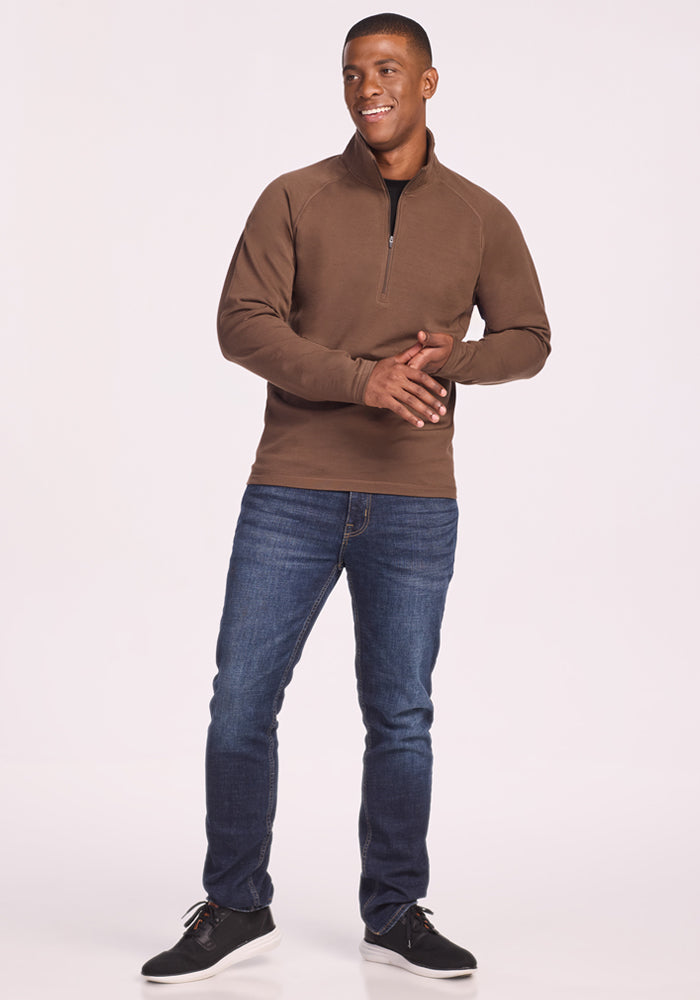 A man stands smiling against a light background, wearing the Woolx Blizzard 1/4 Zip in Bourbon Oak, blue jeans, and black sneakers. His hands are clasped together at chest level.