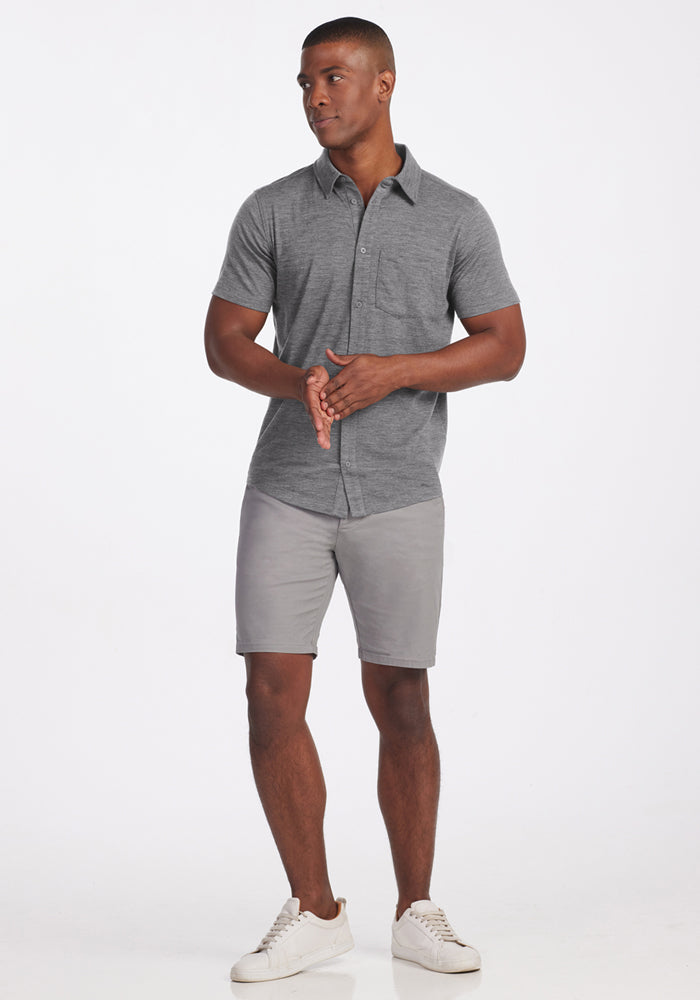 A man stands against a white background wearing the Woolx Endi Full Button Down Shirt in Graphite Heather, paired with light gray shorts and white sneakers. He looks to his right with his hands clasped in front.