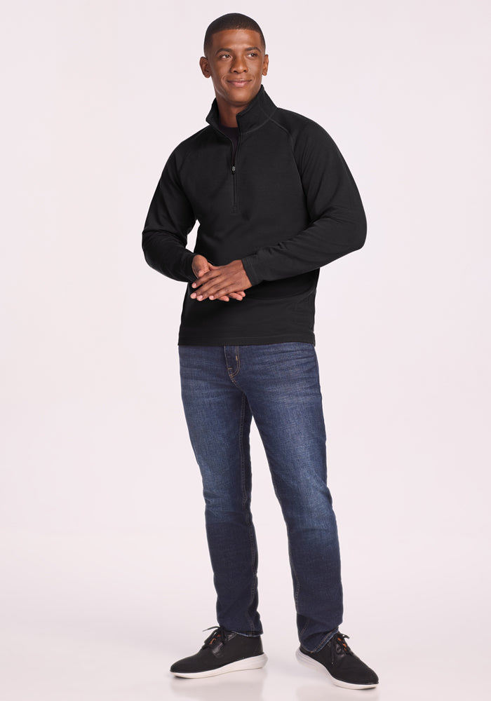 A man stands against a plain background, smiling and clasping his hands while wearing the Woolx Blizzard 1/4 Zip pullover in black, blue jeans, and black shoes.
