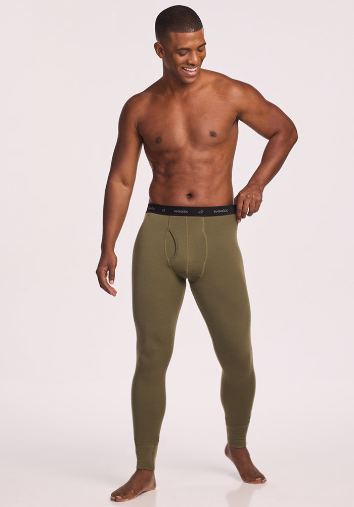 A smiling, shirtless man stands barefoot wearing Woolx Backcountry Leggings in Winter Moss with a black waistband, posing against a plain light background.