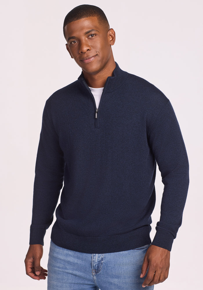A man in the Woolx Enzo Quarter Zip - Storm Cloud sweater over a white t-shirt and light blue jeans stands against a plain light background, looking at the camera with a slight smile. 