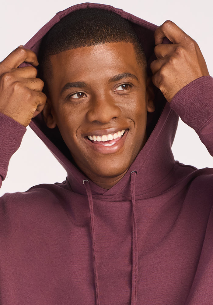 A smiling man in a Woolx Chase Hoodie - Grape Harvest lifts the hood over his head while looking slightly to the side.