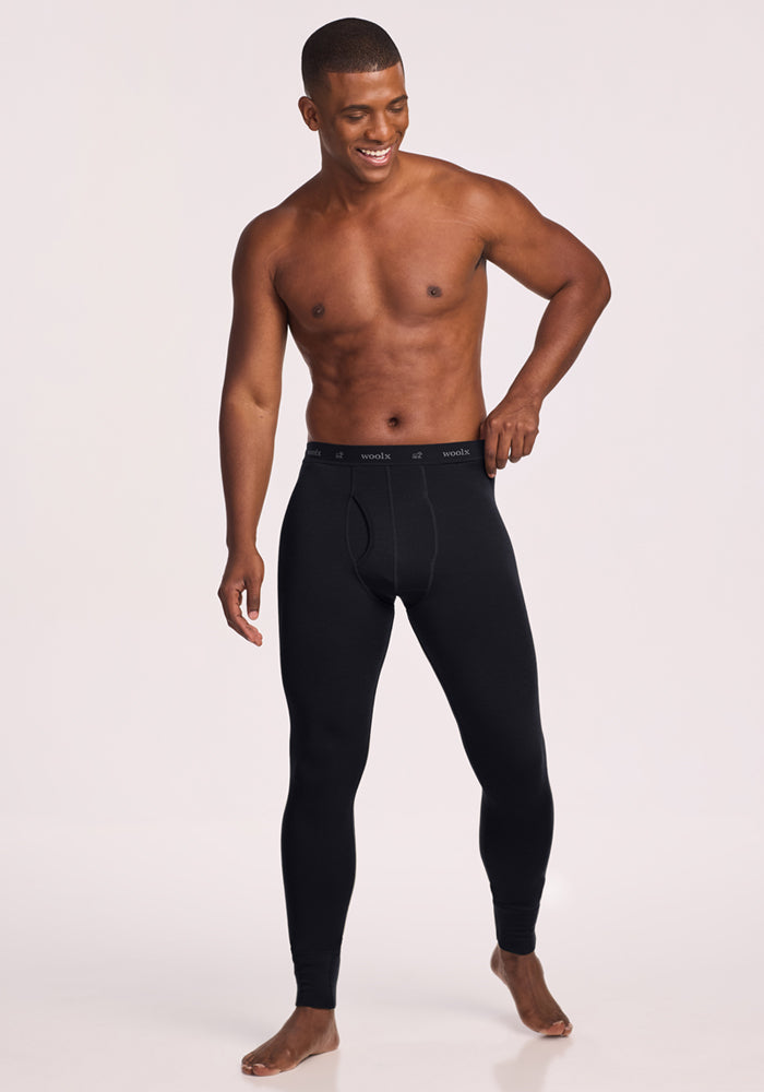 A man stands barefoot against a plain background, wearing only Woolx Backcountry Leggings in black—ideal as a base layer for cold weather adventures.