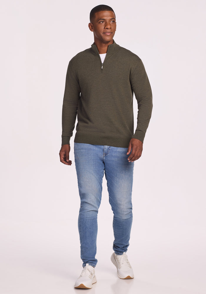 A man wearing a Woolx Enzo Quarter Zip in Pine Needle, light blue jeans, and white sneakers walks forward against a plain light background.