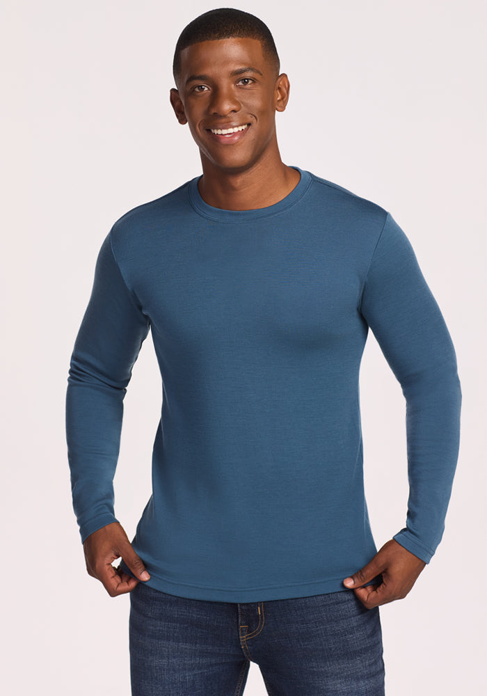A man wearing the Woolx Easton Crew Neck Shirt in steel blue and jeans stands against a plain light background, smiling as he holds the sides of his shirt. 