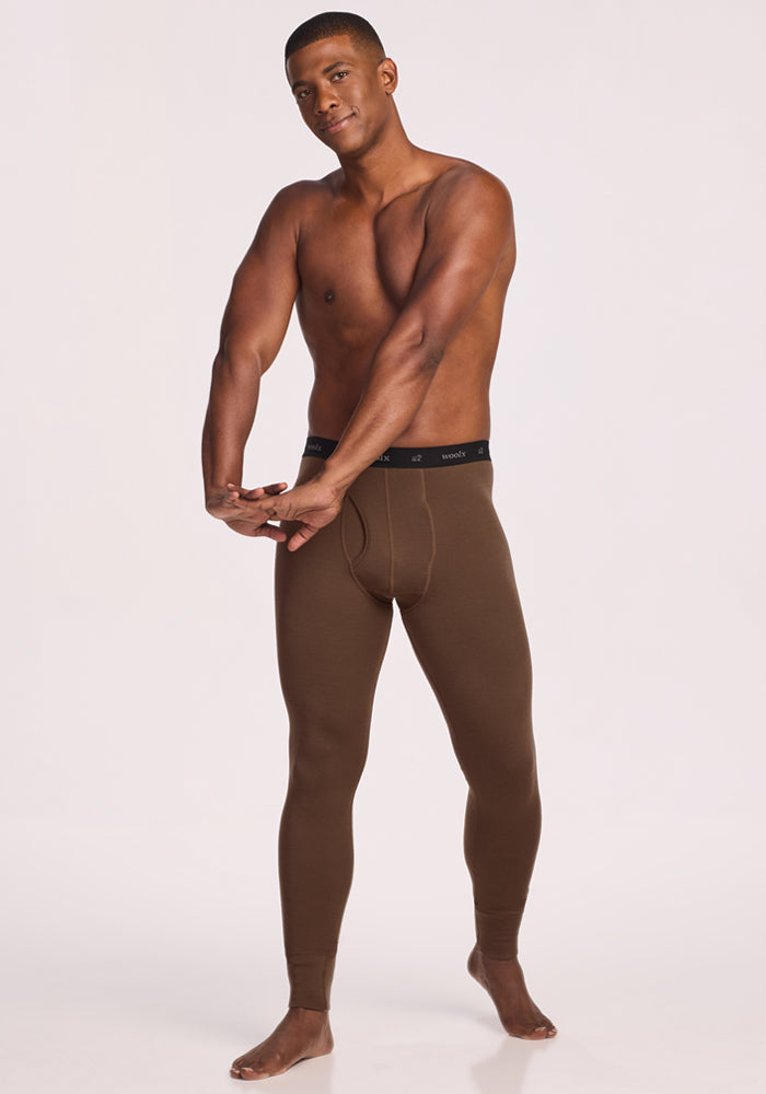 A man stands barefoot against a plain light background, stretching his arms out while wearing Woolx Backcountry Leggings - Bourbon Oak and no shirt, facing the camera.