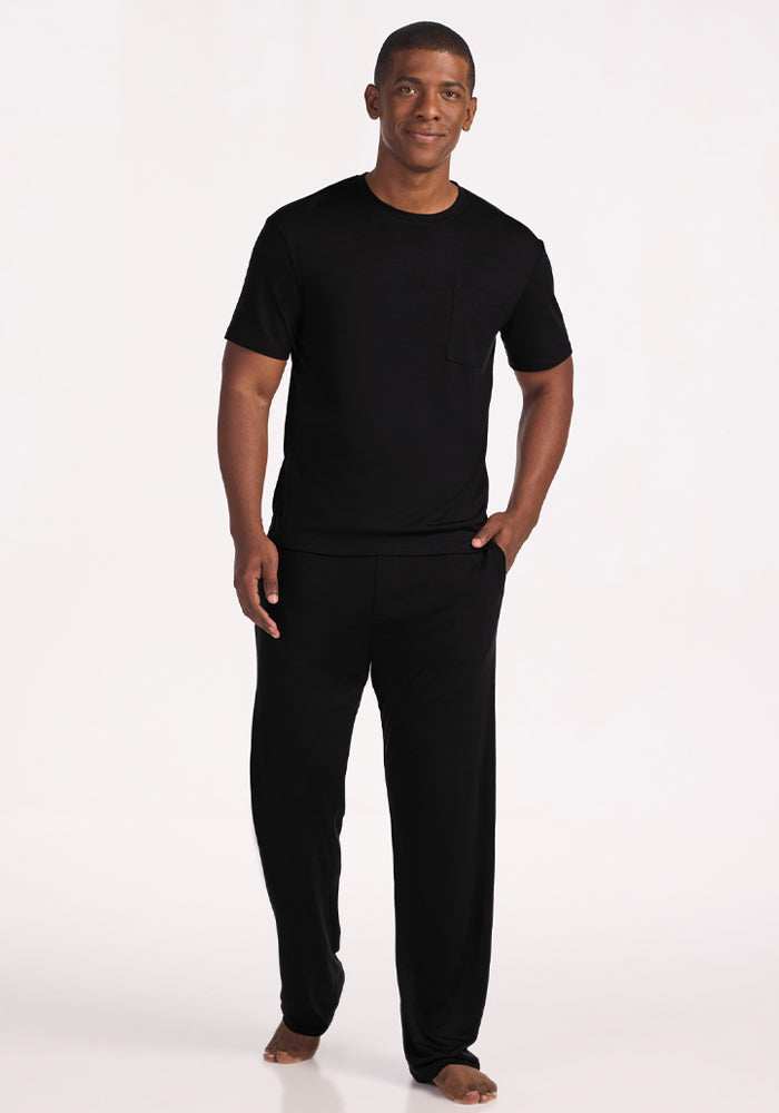 A man stands barefoot against a plain light background, smiling in the Woolx Finn Top - Black, a Merino wool sleep tee with a chest pocket—moisture-managing and odor-resistant for comfort. One hand rests casually in his pocket.