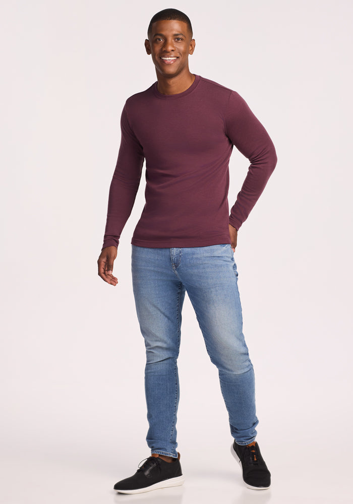 A man smiles against a plain white background, wearing the Woolx Easton Crew Neck Shirt in Grape Harvest, light blue jeans, and black sneakers.