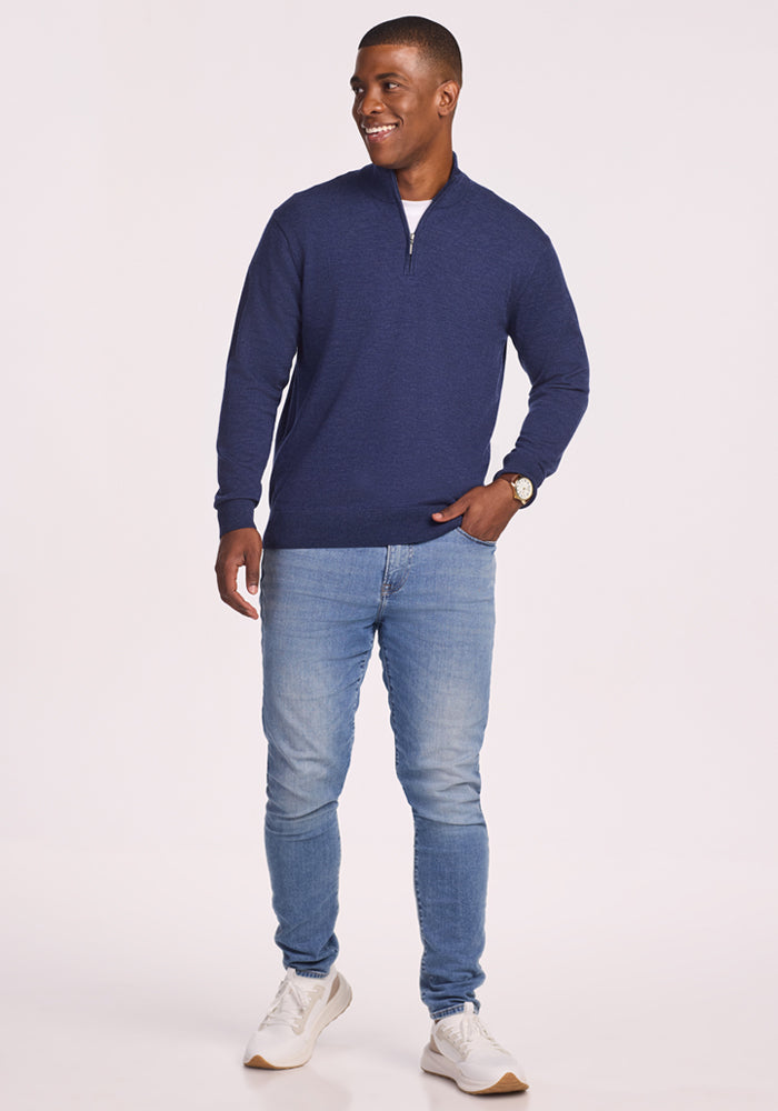 A man smiles against a plain background, wearing the Woolx Enzo Quarter Zip - Twilight Storm sweater with light blue jeans, white sneakers, and a watch. One hand is in his pocket; the other rests at his side.
