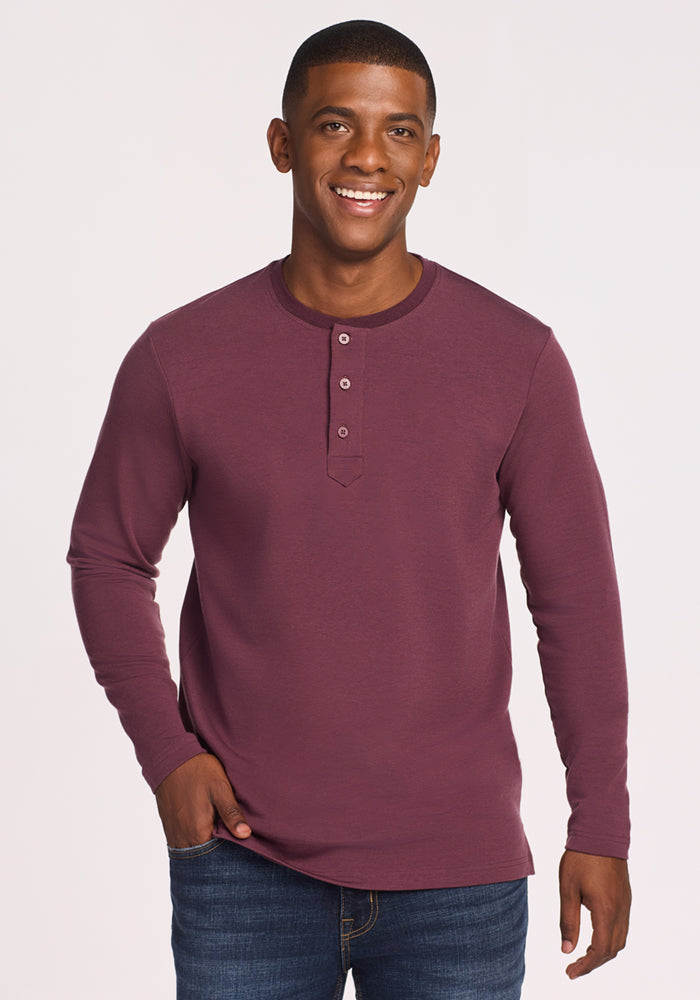 A man wearing the Woolx Jackson Henley in Grape Harvest and blue jeans smiles at the camera with one hand in his pocket, standing against a plain, light background. 