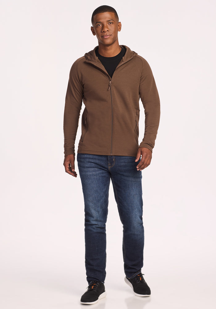 A man stands facing forward wearing the Woolx Boulder Hoodie in Bourbon Oak, a black shirt, blue jeans, and black sneakers against a plain white background.
