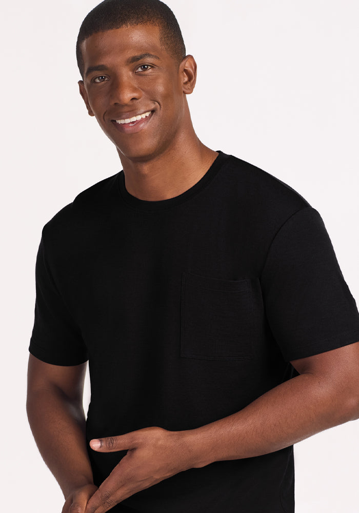 A man with short hair wears the Woolx Finn Top - Black, an odor-resistant Merino wool sleep tee, standing against a light background, smiling gently and gesturing with one hand.