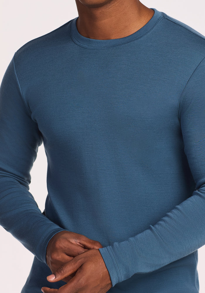A person is shown from shoulders to waist wearing the Woolx Easton Crew Neck Shirt in steel blue, a fitted, moisture-wicking long sleeve shirt, with hands clasped at the torso.