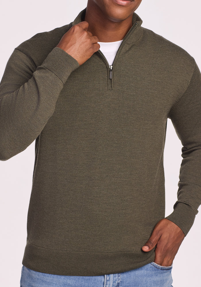 A person wears the Woolx Enzo Quarter Zip in Pine Needle over a white shirt and blue jeans, one hand adjusting the zipper and the other on their hip. The cropped image focuses on their upper body.