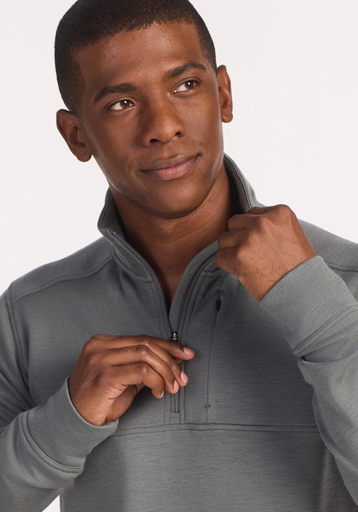 A man wearing the Woolx Mason 1/4 Zip in Sedona Sage looks slightly to the side and smiles softly while adjusting the zipper with one hand.