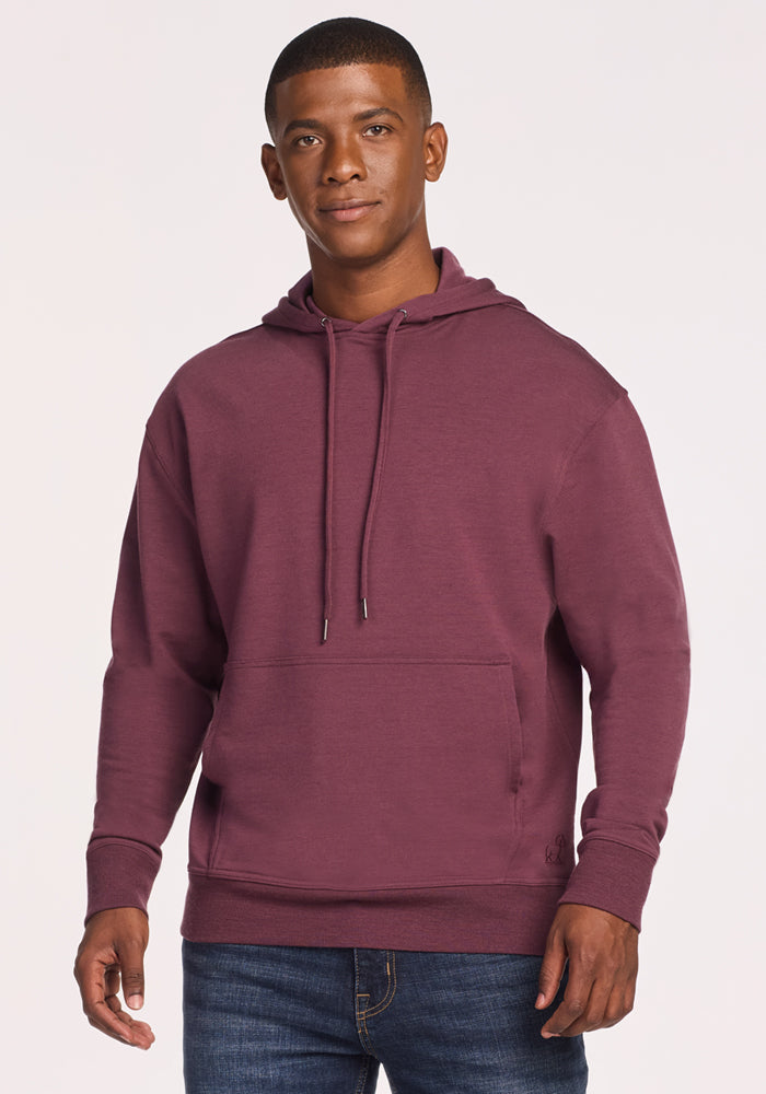 A man wearing the Woolx Chase Hoodie in Grape Harvest and blue jeans stands against a plain light background, looking at the camera with a neutral expression. 