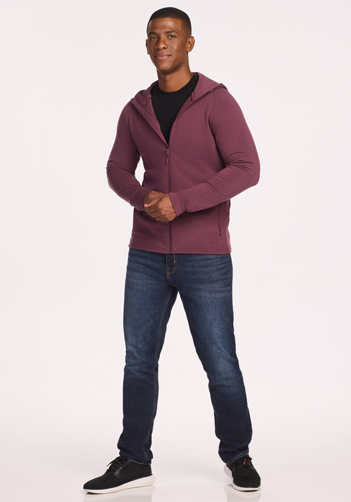A man faces forward against a white background, wearing the Woolx Boulder Hoodie - Grape Harvest over a black shirt, blue jeans, and black sneakers.