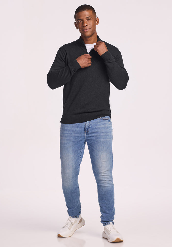 A man stands against a white background, wearing the Woolx Enzo Quarter Zip - Coal Stone sweater over a white shirt, light blue jeans, and white sneakers. He holds the collar of his midweight sweater and looks toward the camera.