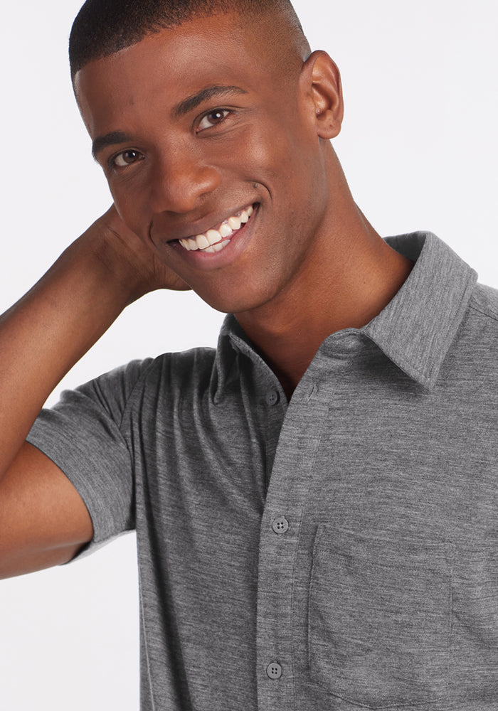 A smiling man stands against a plain light background, wearing the Woolx Endi Full Button Down Shirt in Graphite Heather, with one hand resting behind his neck.
